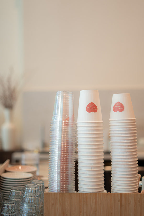 Stacks of clear plastic cups and white paper cups with Rise Bakehouse logos on a wooden surface.
